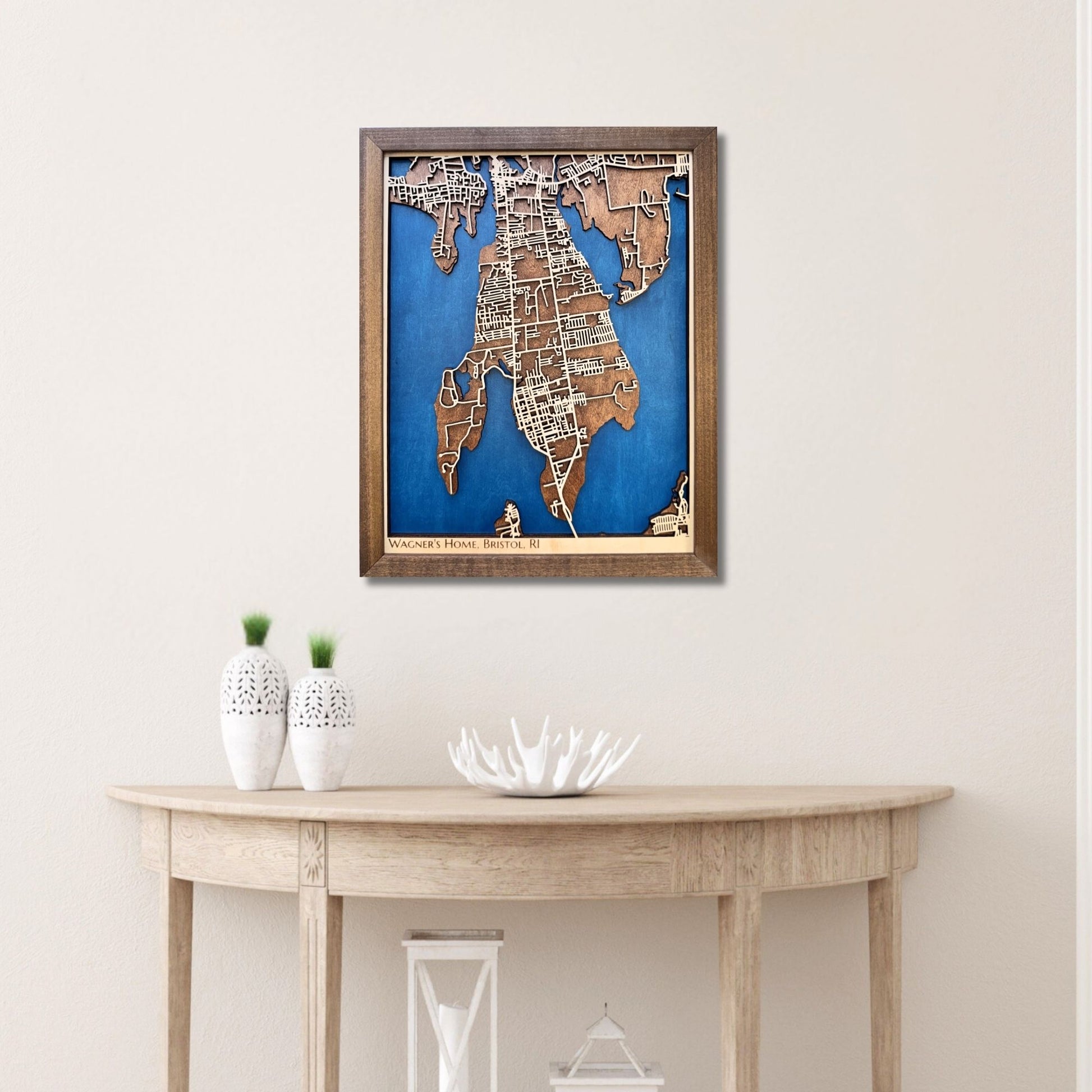 Framed wooden map of Bristol, RI hanging on wall above a wooden table with decorative items. 