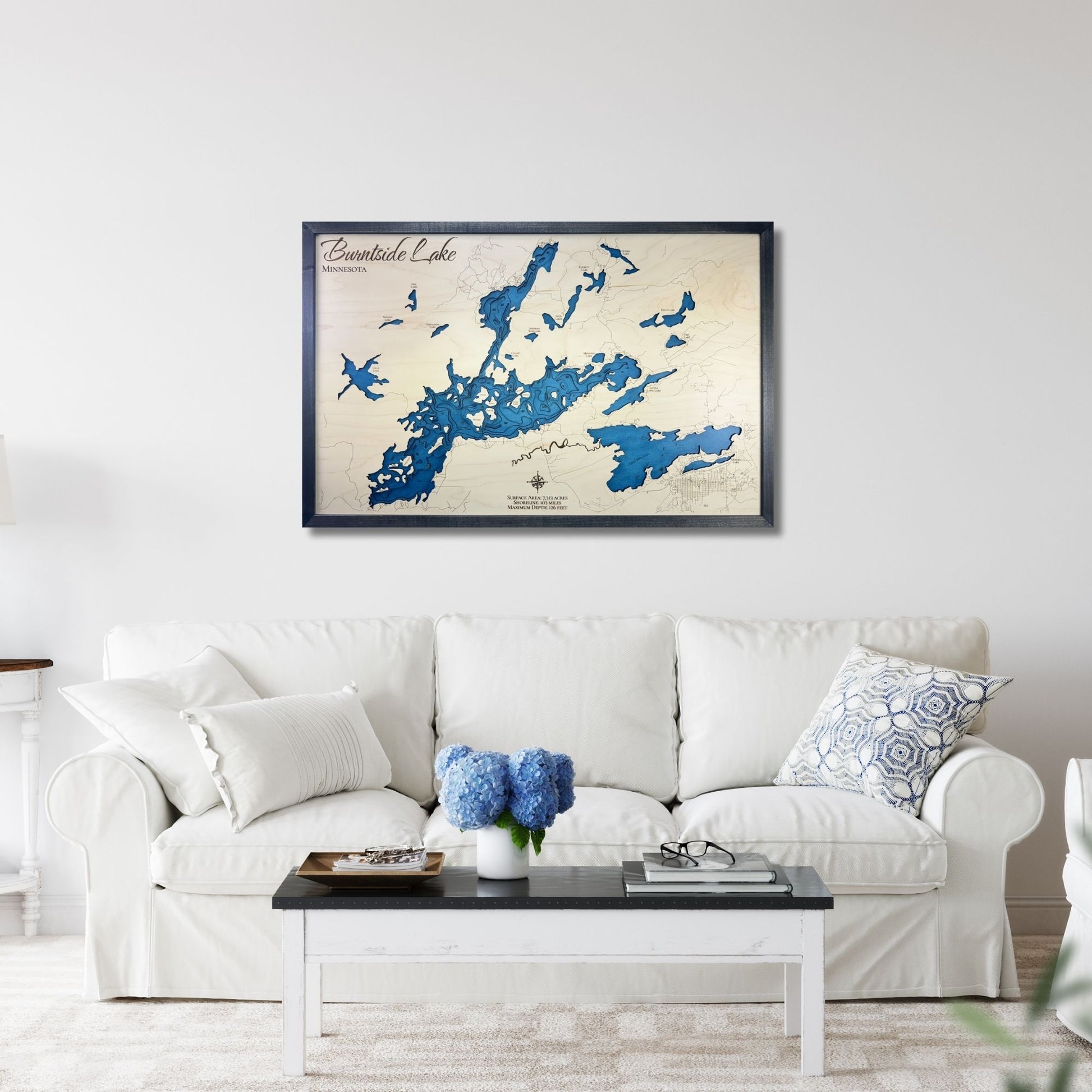 Framed wooden map of Burntside Lake on a wall above a white sofa with decorative pillows.