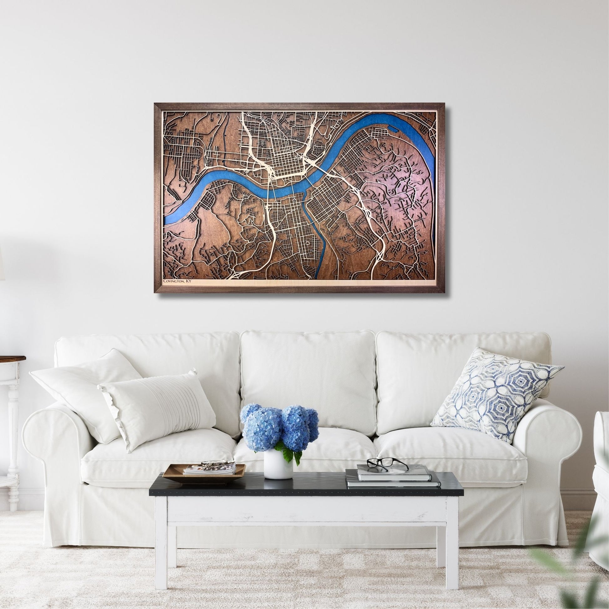3D wooden map of Covington, KY hanging on a wall above white sofa. 