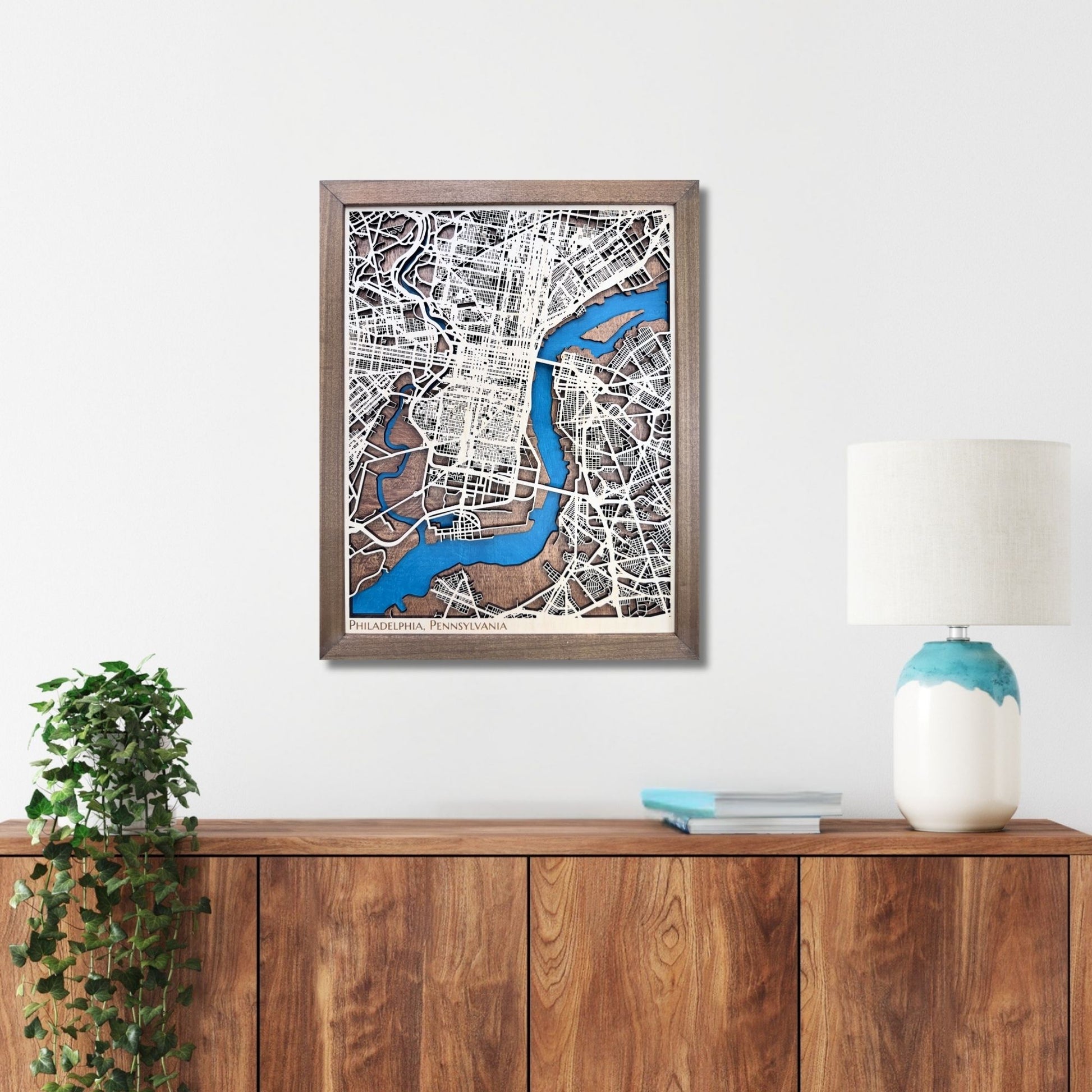 Framed wooden Philadelphia city map on a wall above a wooden console table with a lamp and plant.