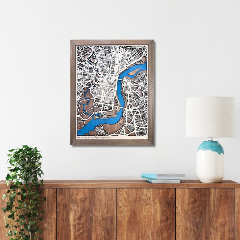 Framed wooden Philadelphia city map on a wall above a wooden console table with a lamp and plant.