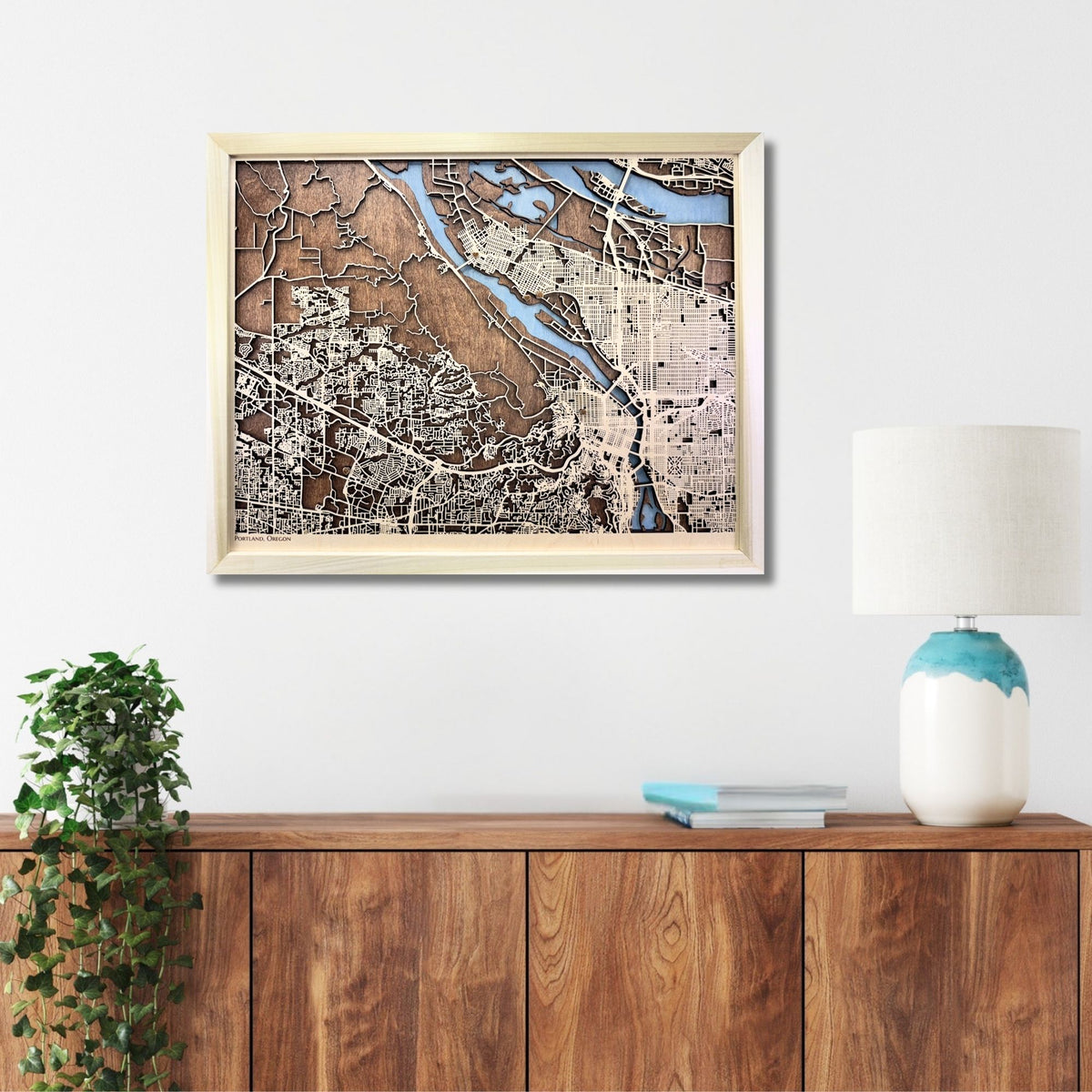 Framed Portland city map on a wall above a wooden cabinet with a plant and lamp.