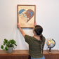 A woman is hanging the two-toned (yin yang) heart-shaped map on a wall above a dresser. 
