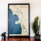Framed wooden map of Pebble Beach Golf Links on a wall with a table and plants below.