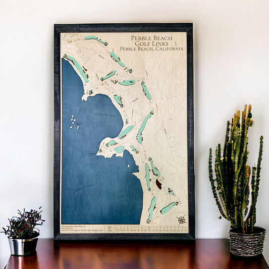 Framed wooden map of Pebble Beach Golf Links on a wall with a table and plants below.