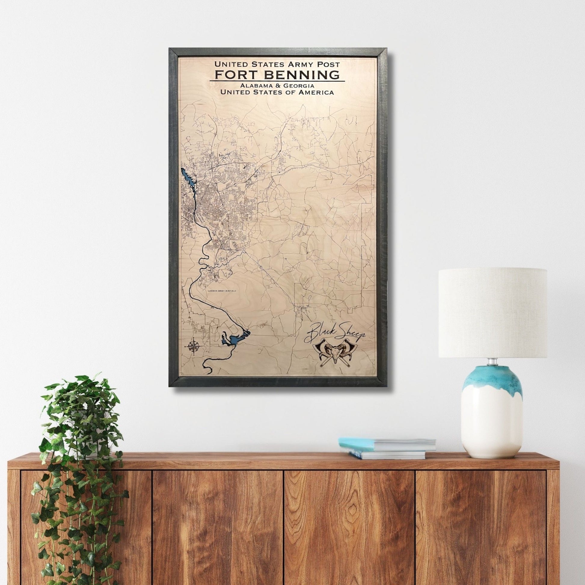 Framed custom wooden military map of Fort Benning on a wall above a wooden sideboard with decor items.